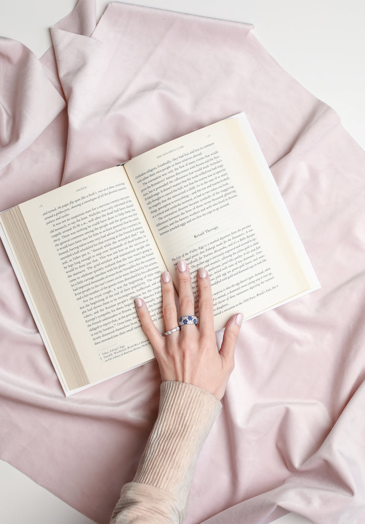 Open book on a pink fabric surface with a hand wearing a ring.
