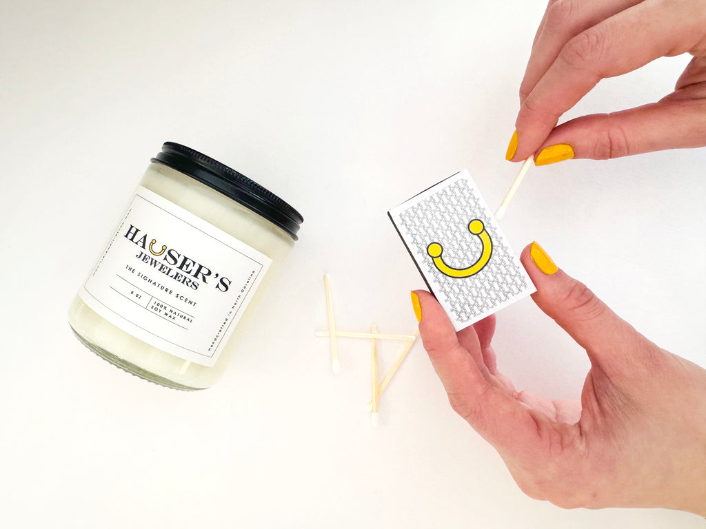 Hauser’s Jewelers Signature Candle in a glass jar with white tea and fig scent, styled with a matchbox and yellow-polished hands